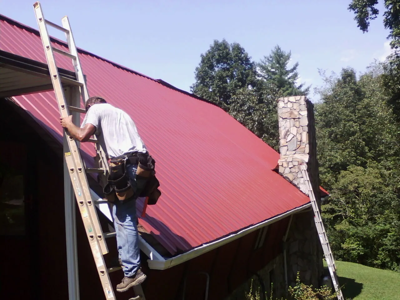 Roofer on a ladder beside a red metal roof and stone chimney — Soffit & Fascia Repair by Roofing Brothers in Paul